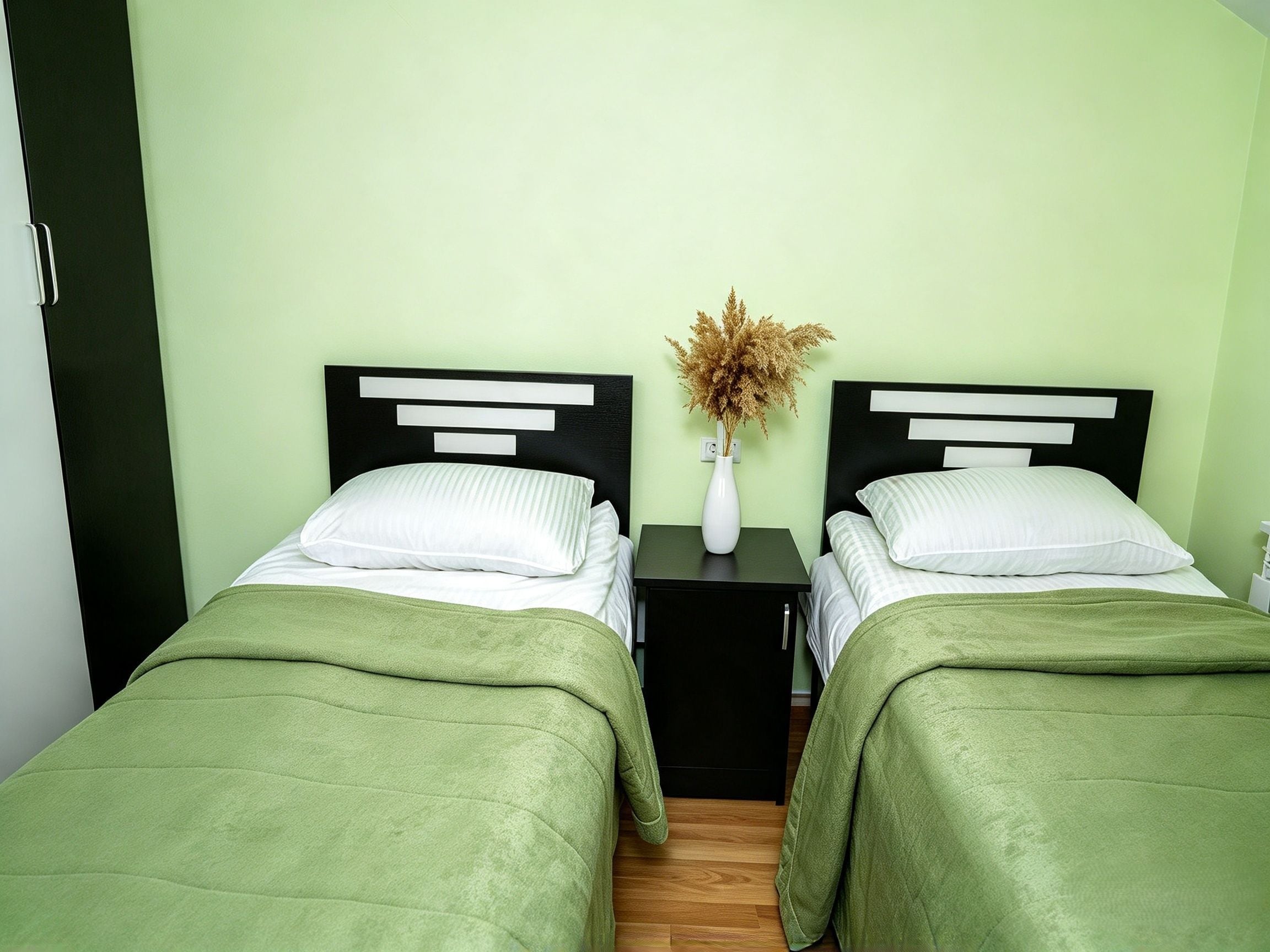 A room with two beds, each with a green bedspread, a white pillow, and a black headboard. There is a small table with a vase of dried plants between the beds.