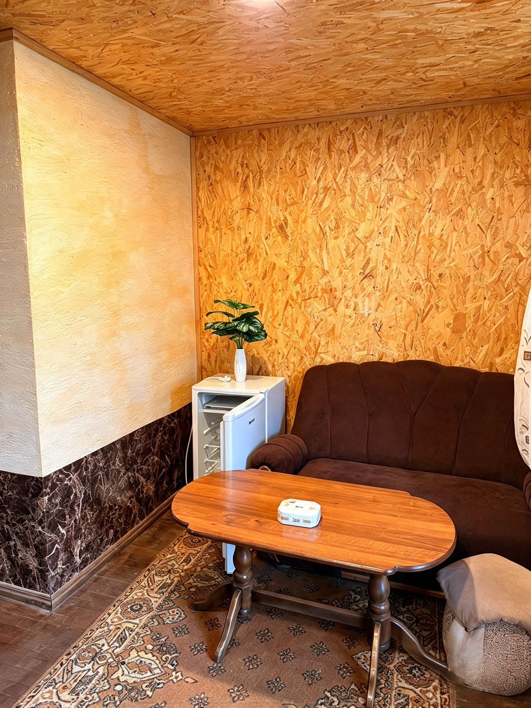 Une pièce avec un plafond en bois, un canapé marron, une petite table sur laquelle est posé un vase, et un tapis à motifs.