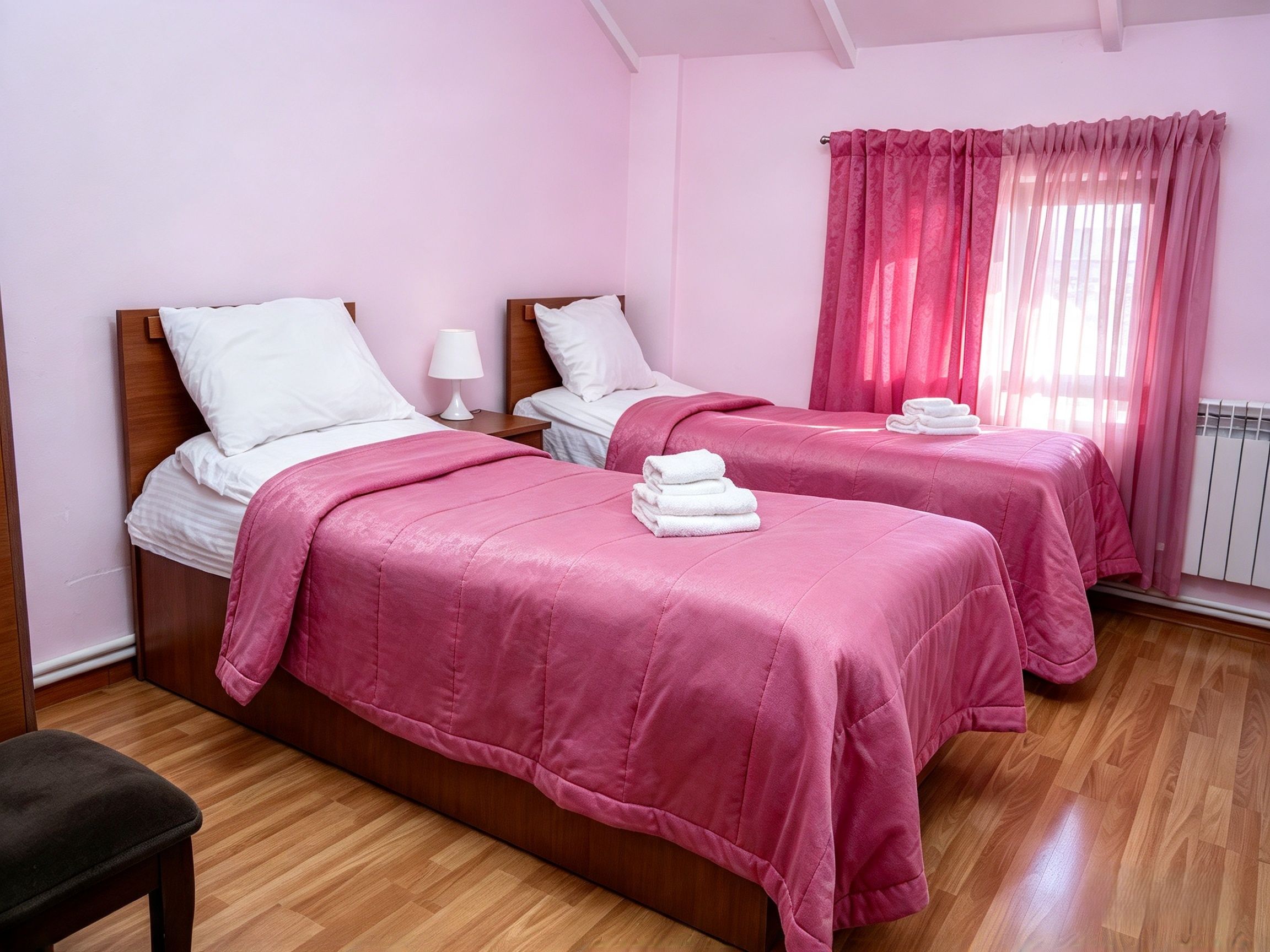 A hotel room with two beds, each with a pink bedspread and white pillows, a wooden floor, and a window with pink curtains.