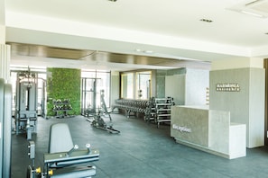 Academia com diversos equipamentos de ginástica, uma mesa de recepção e uma parede com plantas.
