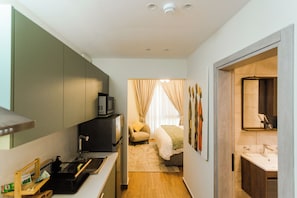 Quarto de hotel compacto com cozinha compacta, cama com colcha verde, pequena área de estar e banheiro com espelho e pia.