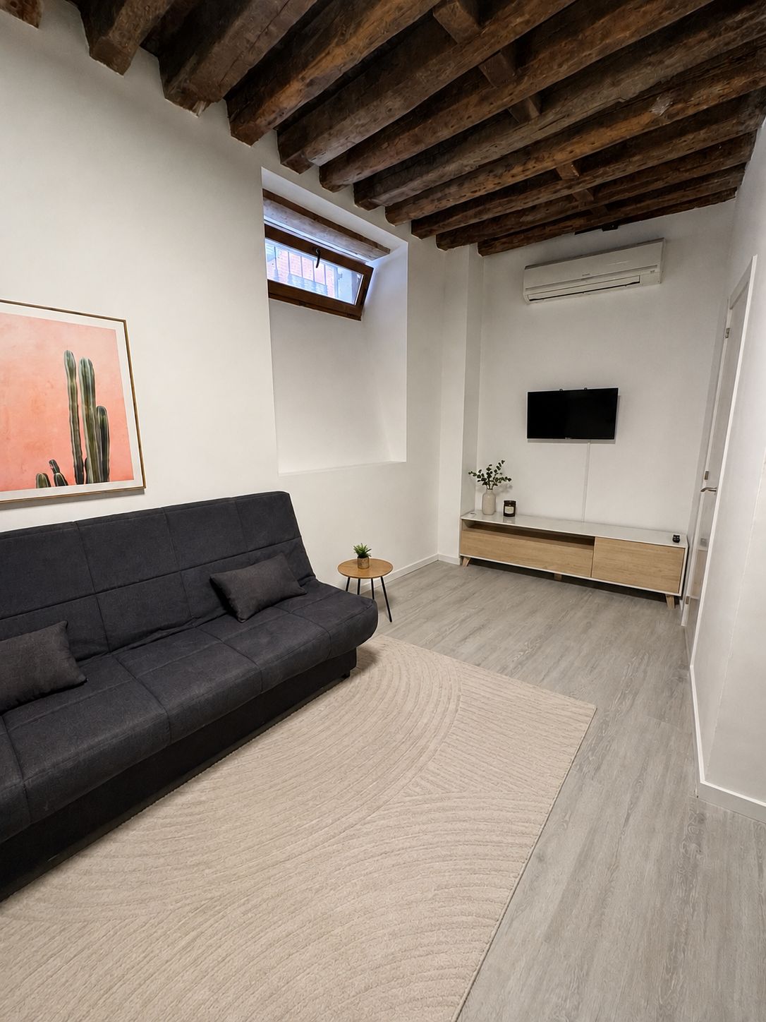 A modern living room with a dark sofa, a small side table, a flat-screen TV mounted on the wall, and a wooden ceiling.