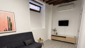 A modern living room with a dark sofa, a small side table, a flat-screen TV mounted on the wall, and a wooden ceiling. - APARTAMENTOS MELISA 12 (Madrid)