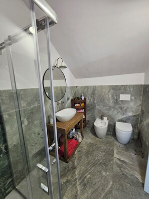 Un baño con cabina de ducha de vidrio, un tocador de madera con lavamanos blanco, un bidé y un inodoro.