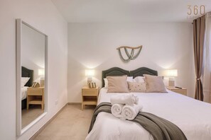 A hotel room with a large mirror, two bedside tables with lamps, a bed with rolled towels, and a wall-mounted light fixture. - Modern 2BR Qala Gozo Apt with Shared Pools (Qala)