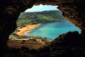 A coastal landscape viewed through a cave opening, featuring a beach, clear blue waters, and distant buildings. - Modern 2BR Qala Gozo Apt with Shared Pools (Qala)