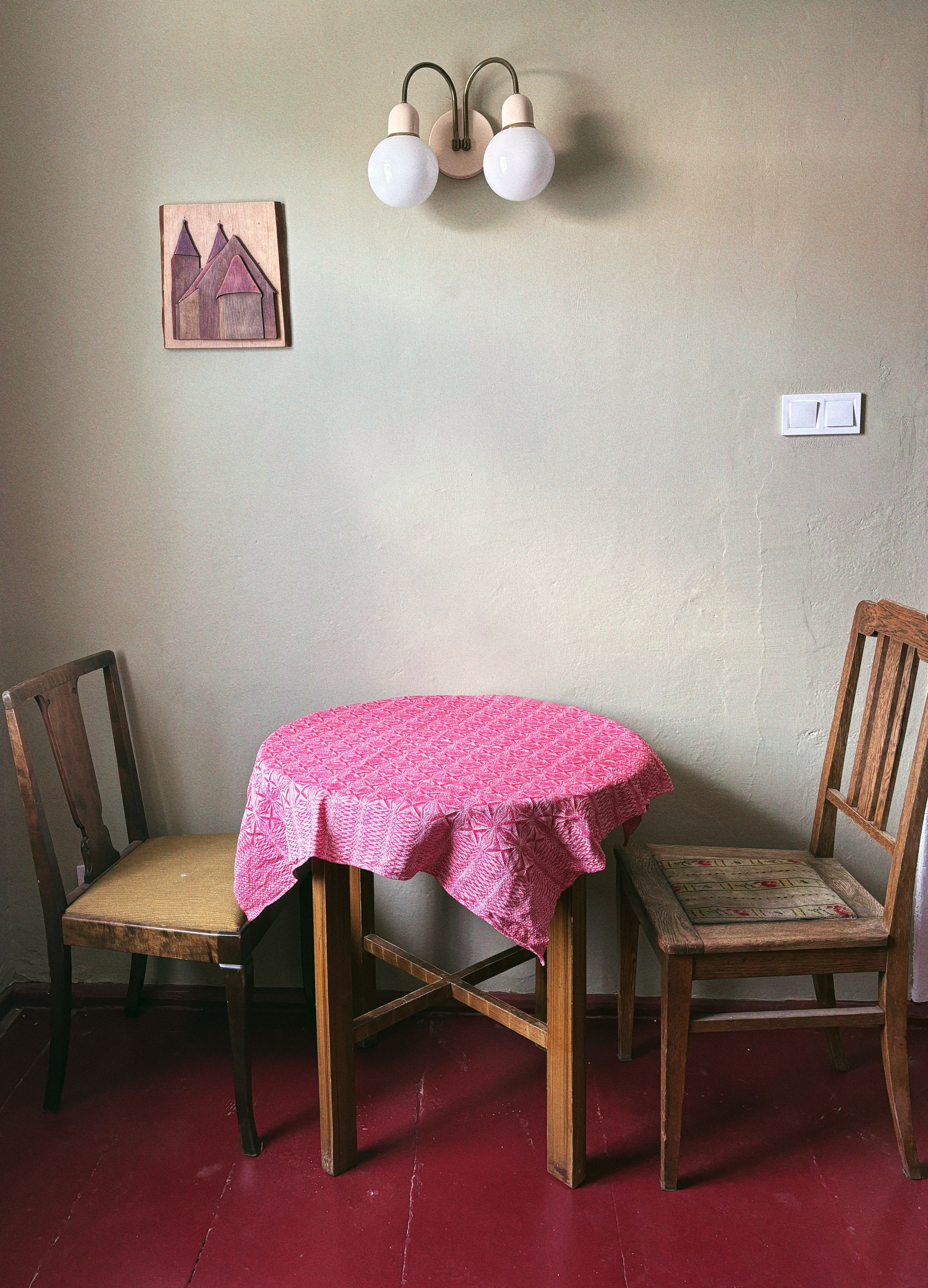 Ein Essbereich mit einem runden Tisch, der mit einer gemusterten Tischdecke bedeckt ist, zwei Holzstühlen, einer wandmontierten Lampe und einem kleinen gerahmten Bild.