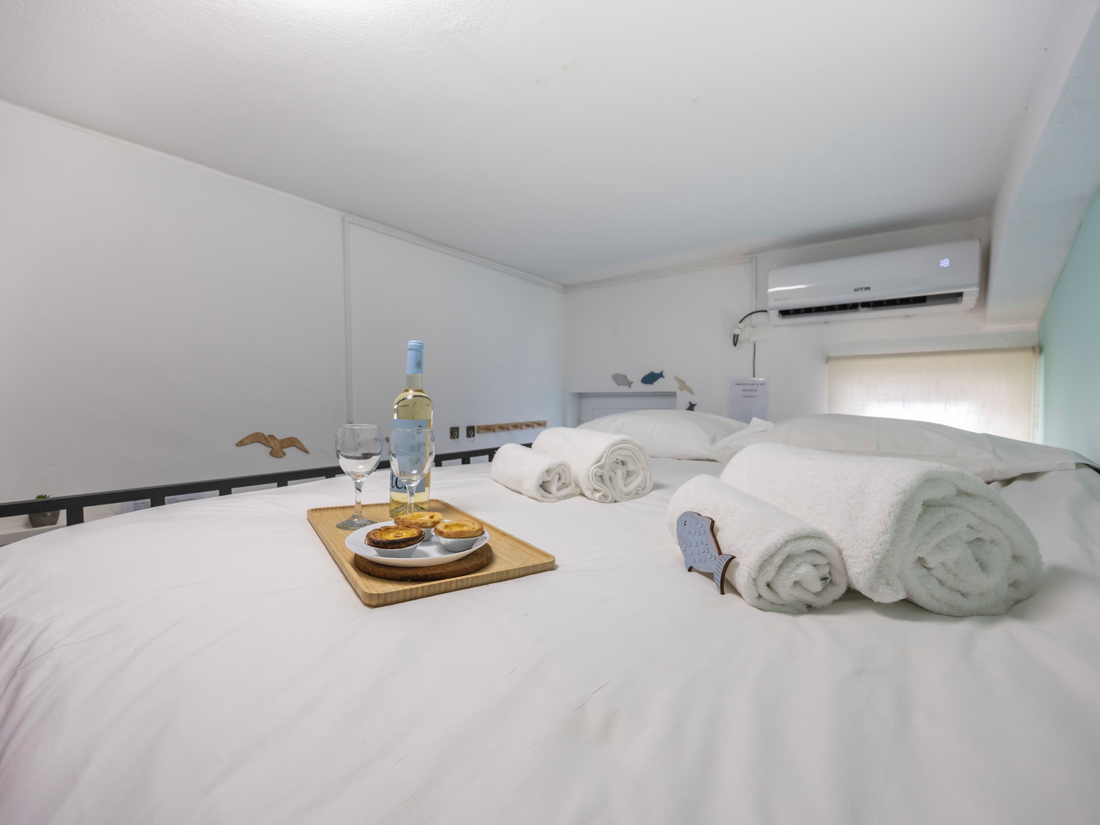 A hotel room with a neatly made bed, a tray with a bottle and glasses, and rolled towels.