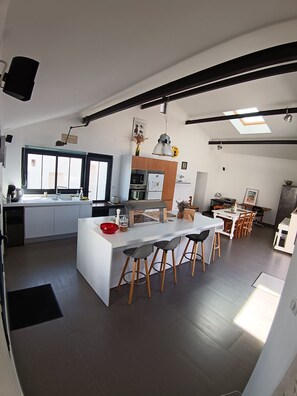 A modern kitchen with a central island, dining area with chairs, and a large window providing natural light. - La Maison de Jeanne - Maison familiale atypique à Brem sur mer (Brem-sur-Mer)