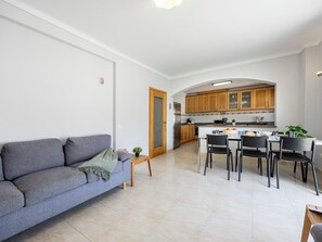Un salon moderne avec un canapé gris, un coin repas comprenant une table et des chaises, et une cuisine dotée de meubles en bois et d’un réfrigérateur en acier inoxydable.