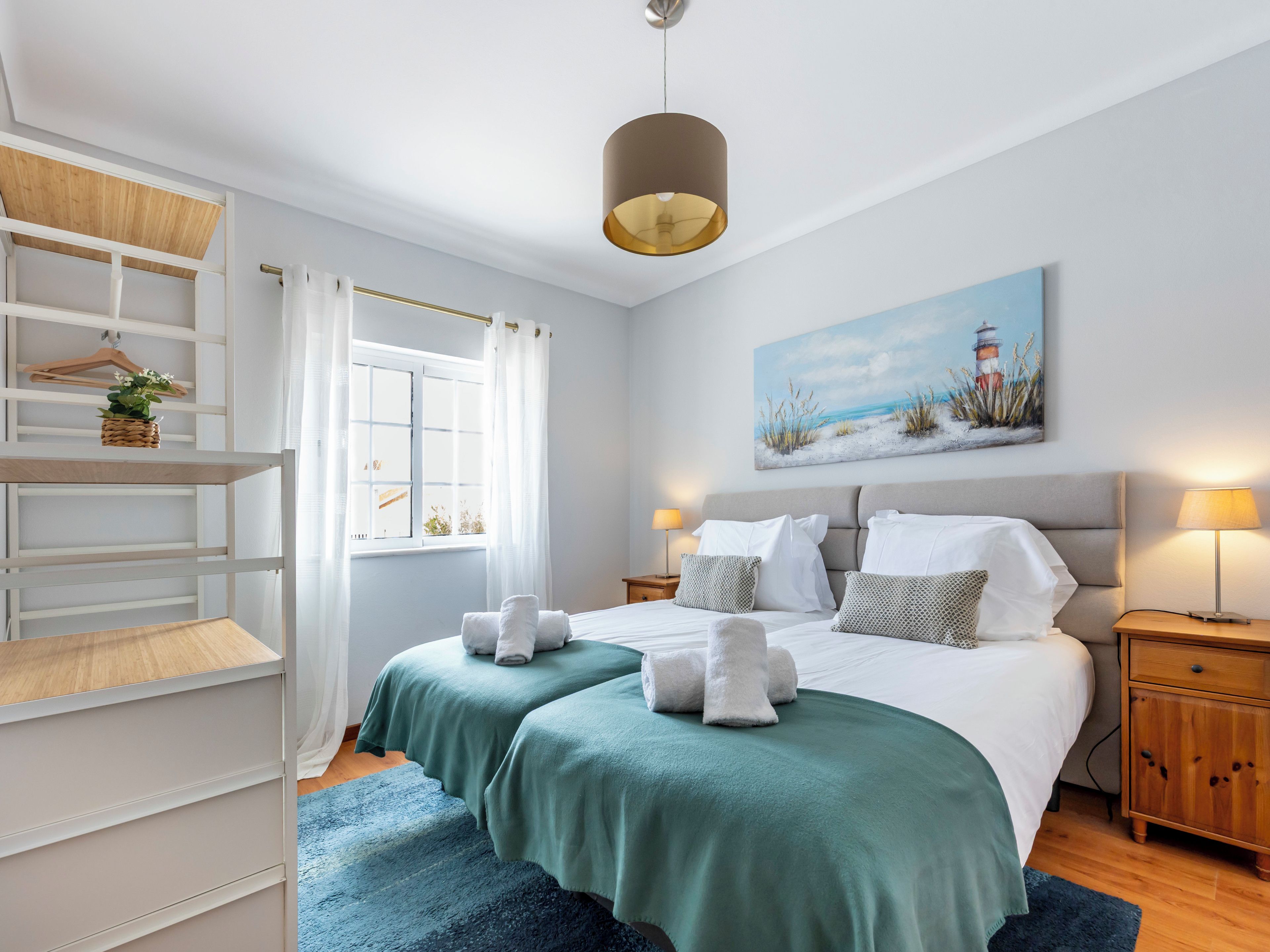 A hotel room with a bed, bedside tables, a wardrobe, a window with curtains, a painting on the wall, and a hanging light fixture.