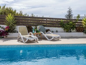 Un espace aménagé au bord de la piscine, avec deux chaises longues, une petite table sur laquelle se trouvent une bouteille et un verre, et une clôture en bois en arrière-plan.