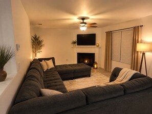A living room with a sectional sofa, a fireplace, a ceiling fan, and a television mounted above the fireplace. - Cozy 3BR Home - Game Days & Family Stays (Tuscaloosa)