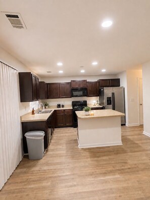 A modern kitchen with wooden cabinets, a stainless steel refrigerator, a microwave, and a central island. - Cozy 3BR Home - Game Days & Family Stays (Tuscaloosa)