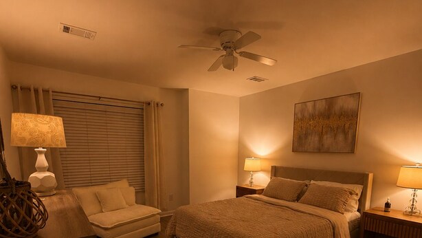 Una camera d'albergo con un letto, una scrivania, una sedia, una ventola a soffitto e un quadro appeso al muro.