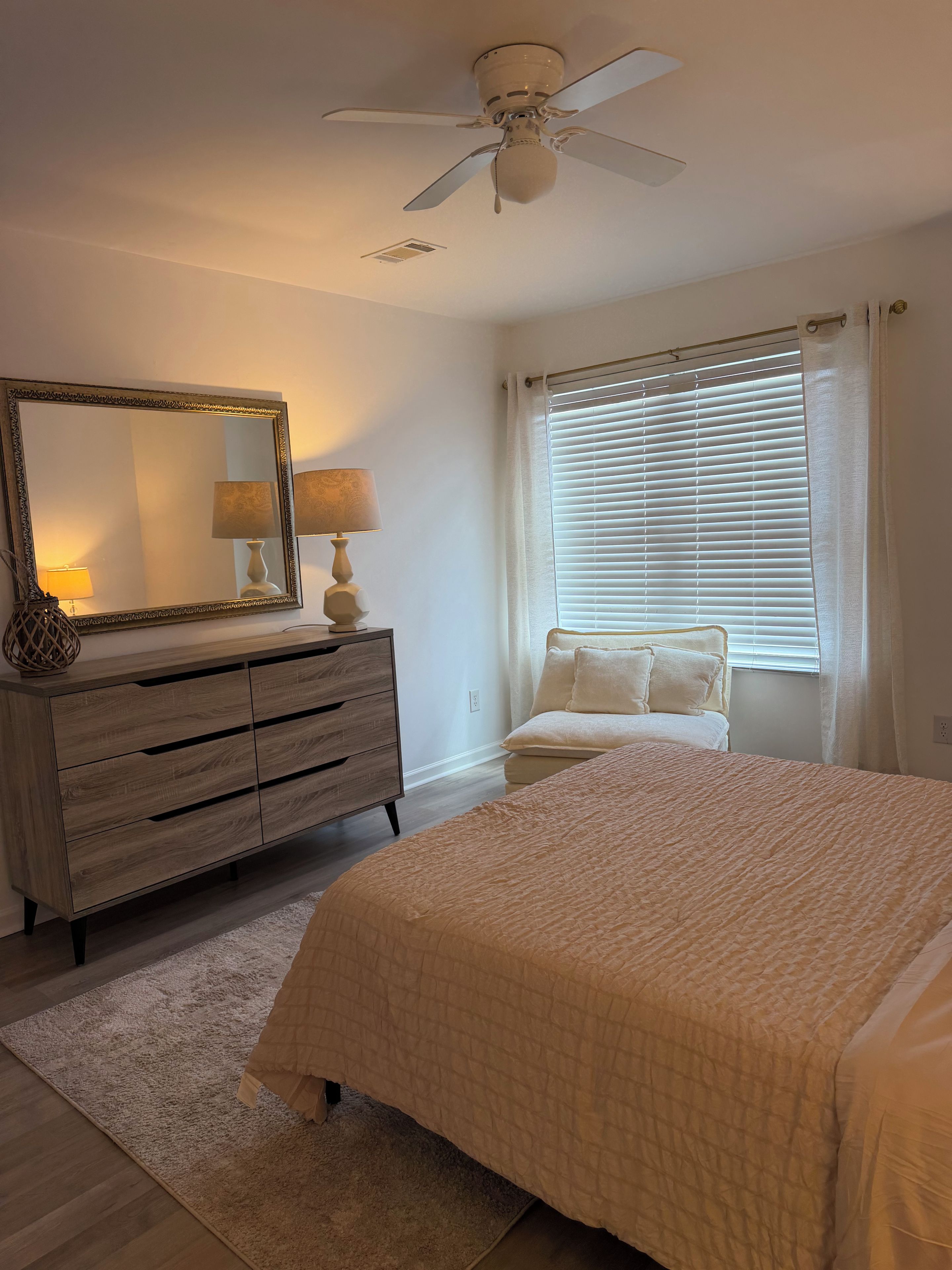 A bedroom with a bed, a dresser, a mirror, a ceiling fan, and a window with blinds.