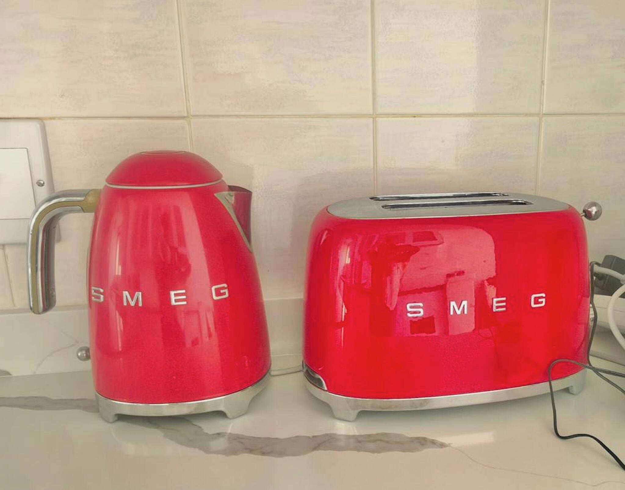 Two red kitchen appliances, a coffee maker and a toaster, with the brand name 'SMEG' on them, placed on a countertop against a tiled wall.