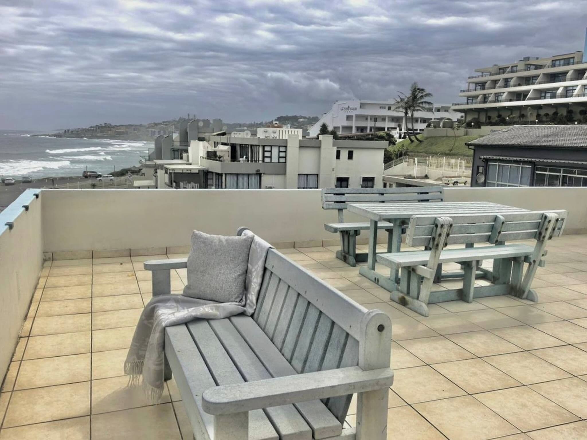 A terrace with a bench and tables, overlooking a coastal area with buildings and the sea.