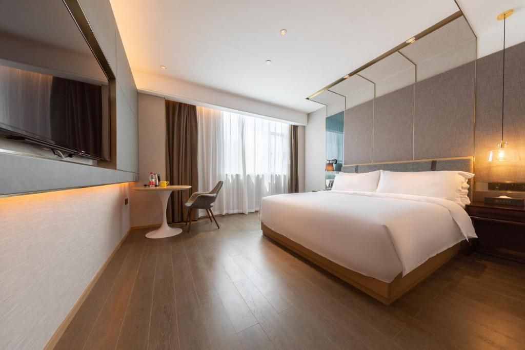 A modern hotel room with a large bed, a flat-screen TV mounted on the wall, a desk with a chair, and a large window with curtains.