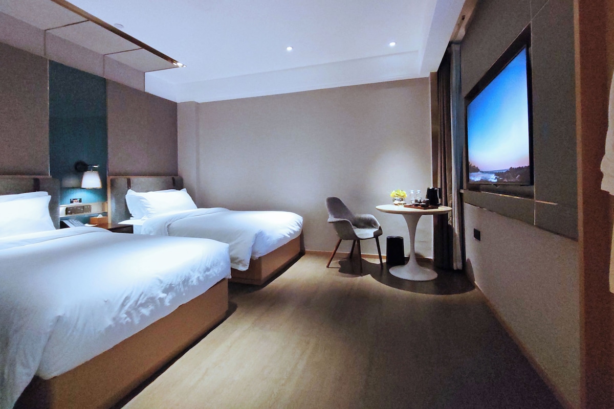 A hotel room with two beds, a desk with a chair, a television, and a small table with a vase and a coffee maker.