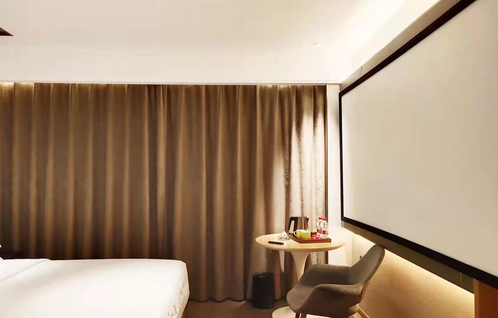 Habitación de hotel con una cama, una mesita con una taza y una bandeja, una silla y una pantalla grande.