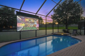 Ein Swimmingpool mit einer großen Leinwand, die einen Filmplakat zeigt, umgeben von einer überdachten Fläche mit Blick auf Bäume und einen Sonnenuntergang.
