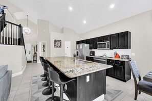 A modern kitchen with a central island, black cabinetry, stainless steel appliances, and a dining area with bar stools. - 6BD Mins to Disney | Pool, Spa, Theater, Game Room (Kissimmee)
