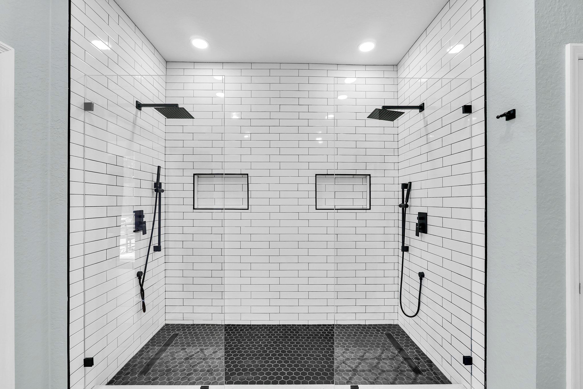 Une salle de douche moderne avec des murs recouverts de carreaux blancs, deux pommes de douche et un sol à motifs hexagonaux.