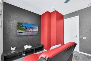 Una habitación con una televisión de pantalla plana montada en una pared texturizada, una consola de videojuegos y un sillón rojo acolchado.