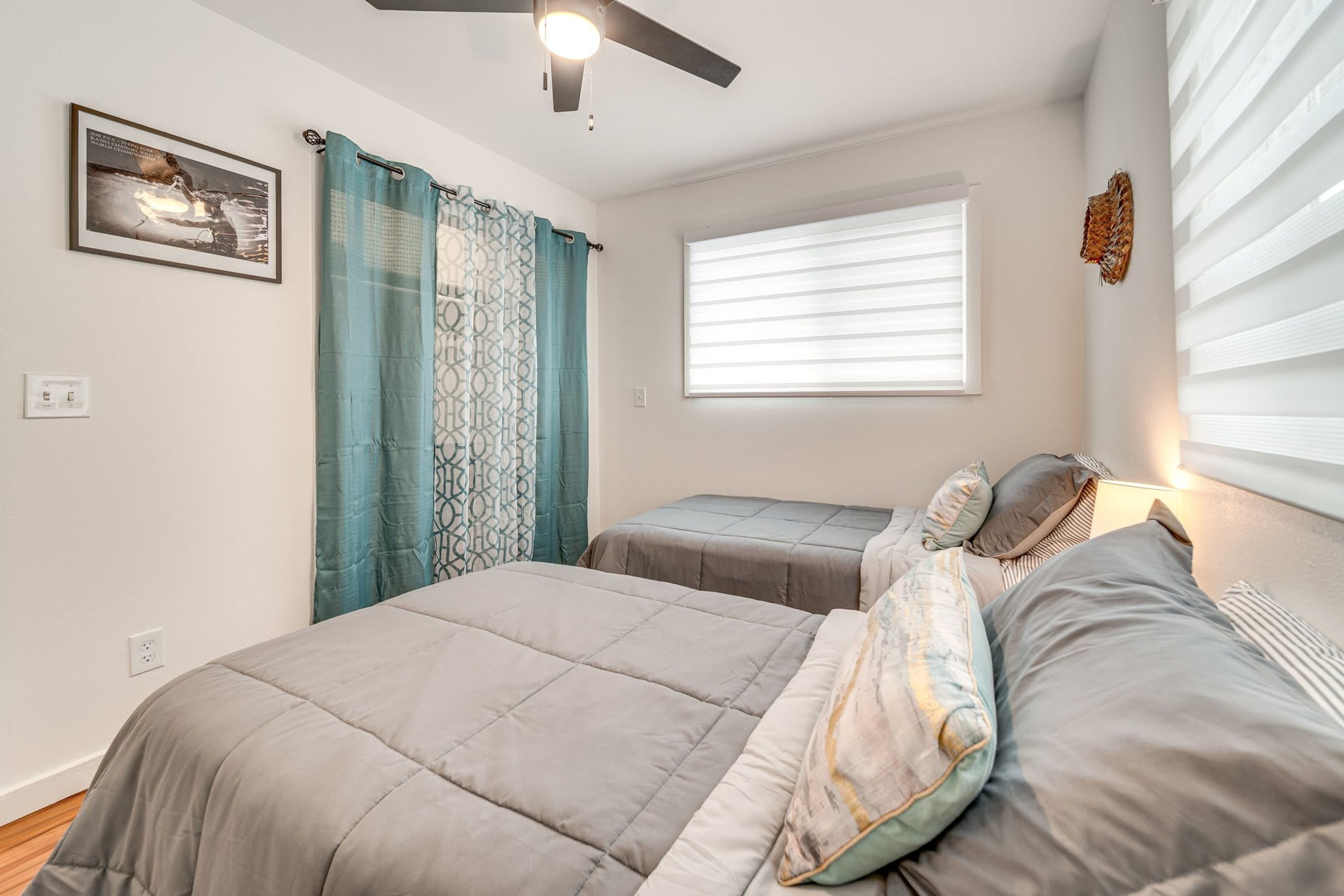 Un dormitorio con dos camas, un ventilador de techo, una ventana con persianas y un cuadro enmarcado en la pared.