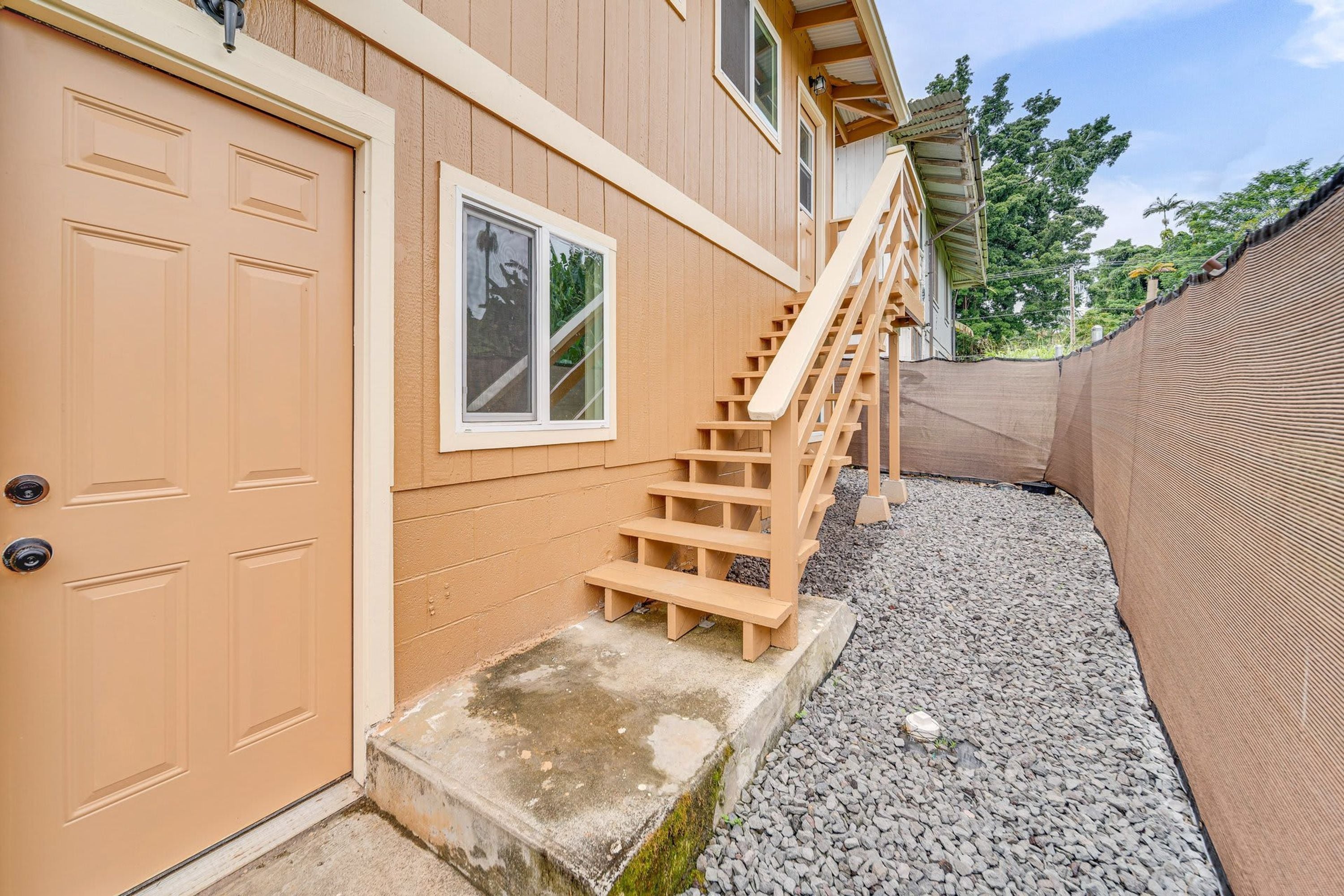 A two-story house with a wooden staircase leading to a front door, a small front porch, and a gravel driveway.
