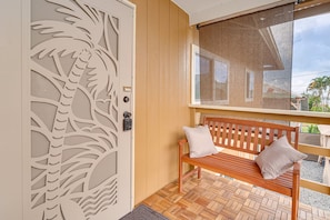 A wooden bench with cushions, a patterned door, and a window with a view of a house and palm trees. - Modern Hilo Hideaway - Gated Yard Gazebo (Hilo)