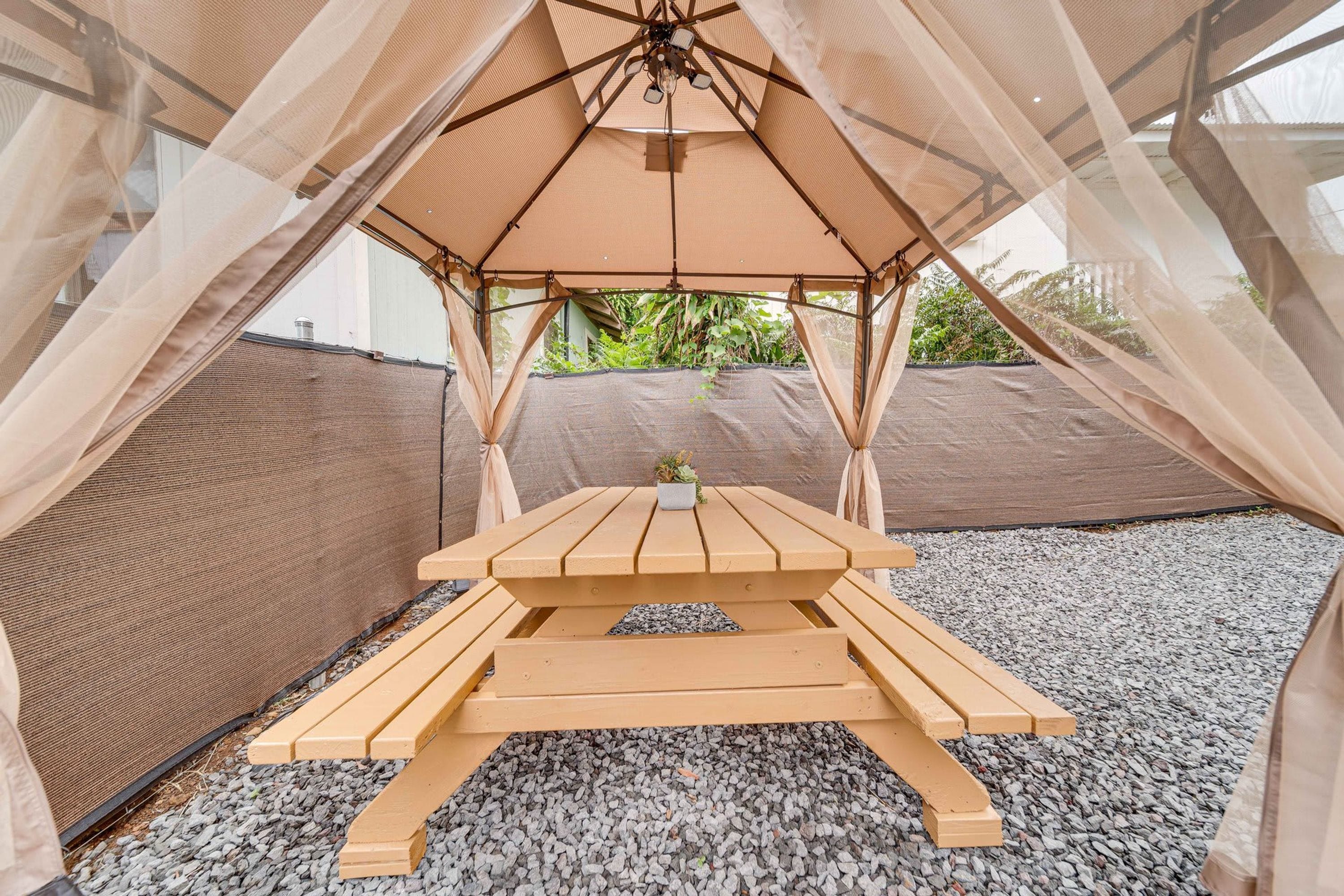 Una mesa de picnic de madera bajo una carpa con toldo de malla, rodeada de grava.