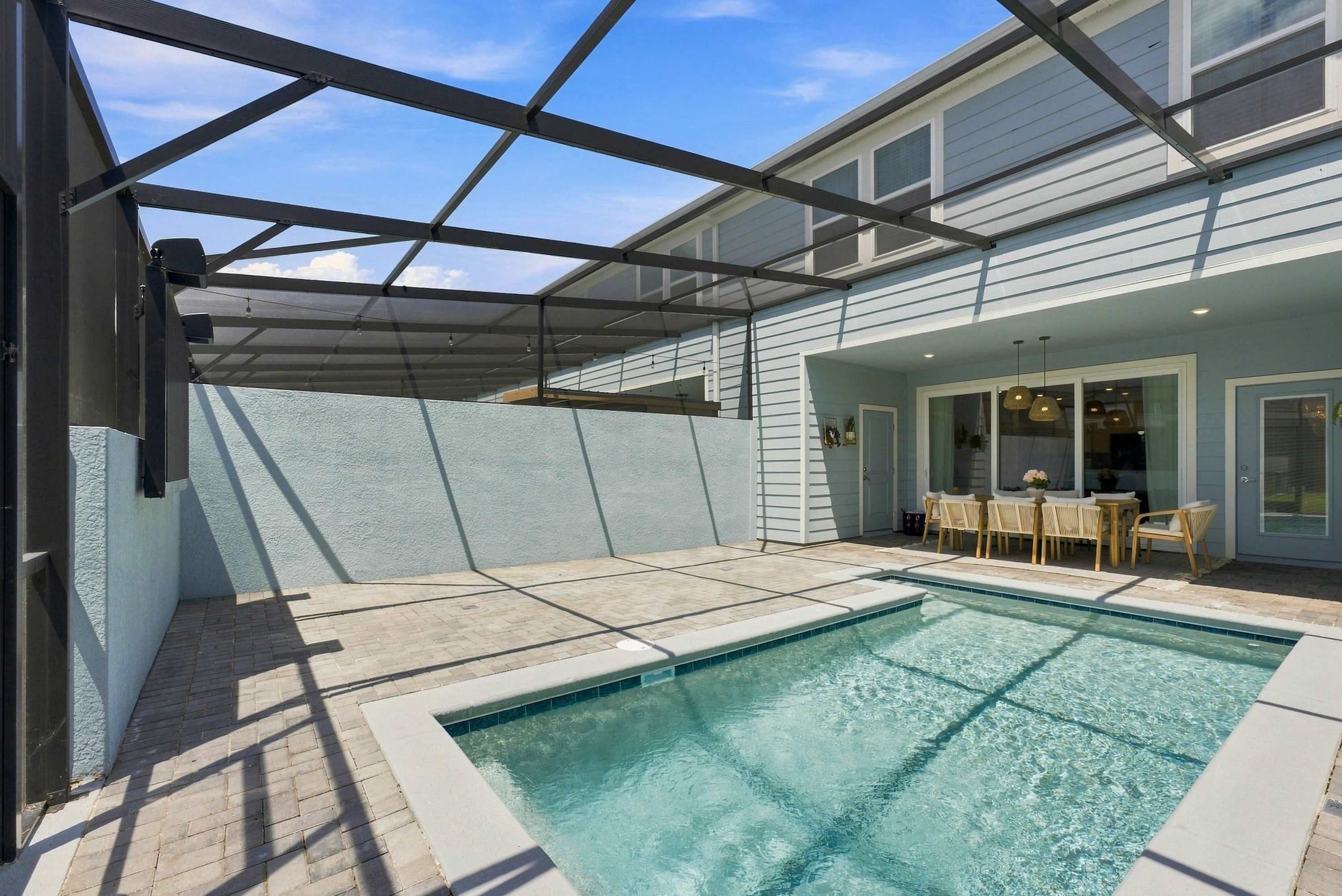Ein Schwimmbad mit überdachter Terrasse, einem Esstisch mit Stühlen und einem Haus mit hellblauem Anstrich.