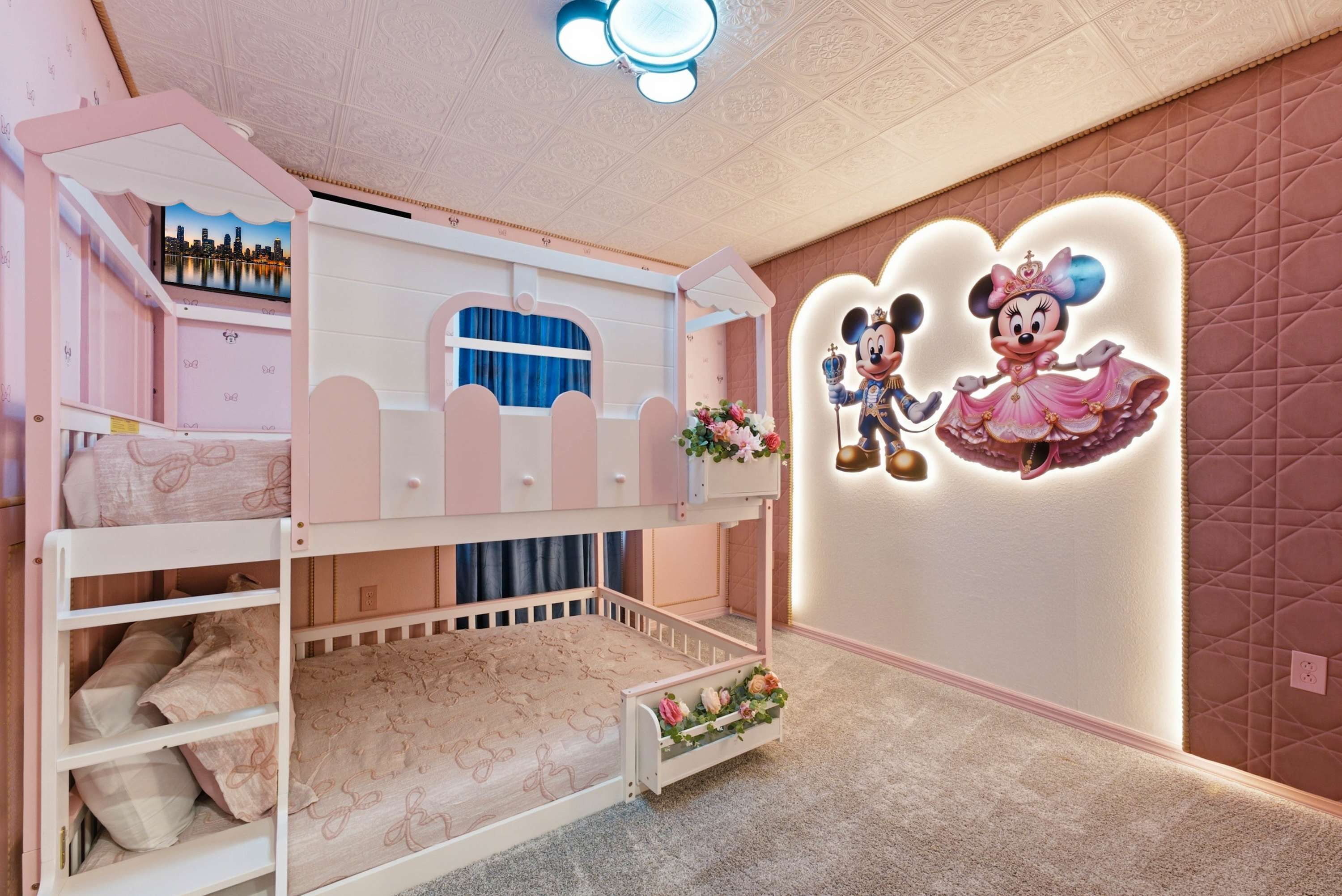 Un lit superposé avec un téléviseur diffusant des images de la ville, une décoration murale à l’effigie de Mickey et Minnie, et un petit arrangement floral.