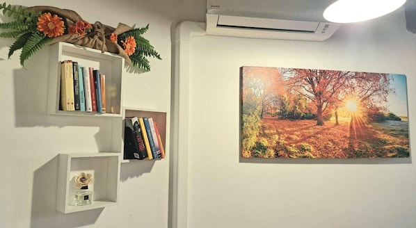 A wall with a framed painting of a sunset, a white shelf with books, and a small vase with a flower.