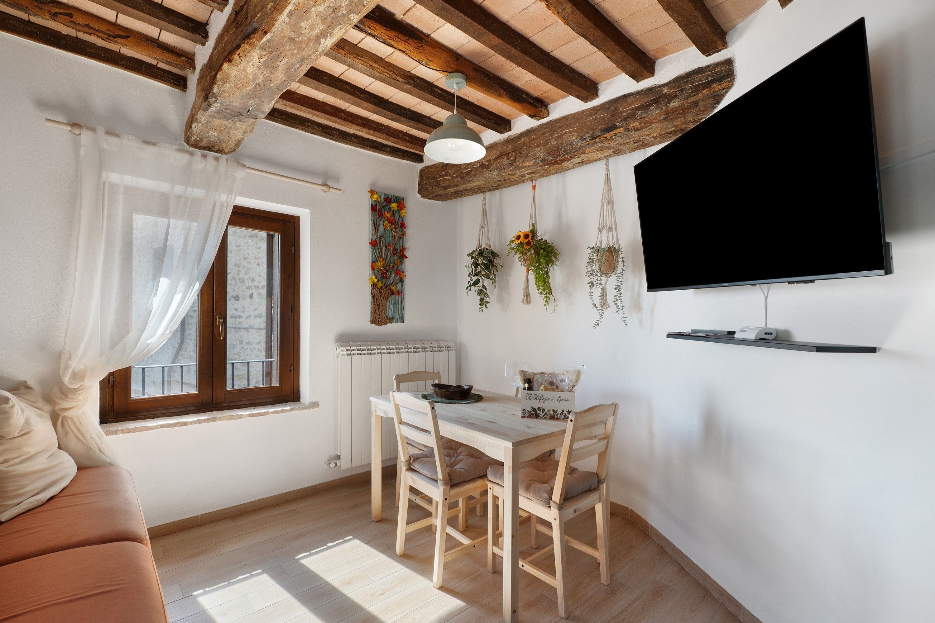 A cozy living room with a wooden ceiling, a dining area with a table and chairs, a television mounted on the wall, and a window with sheer curtains.