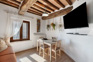 A cozy living room with a wooden ceiling, a dining area with a table and chairs, a television mounted on the wall, and a window with sheer curtains.