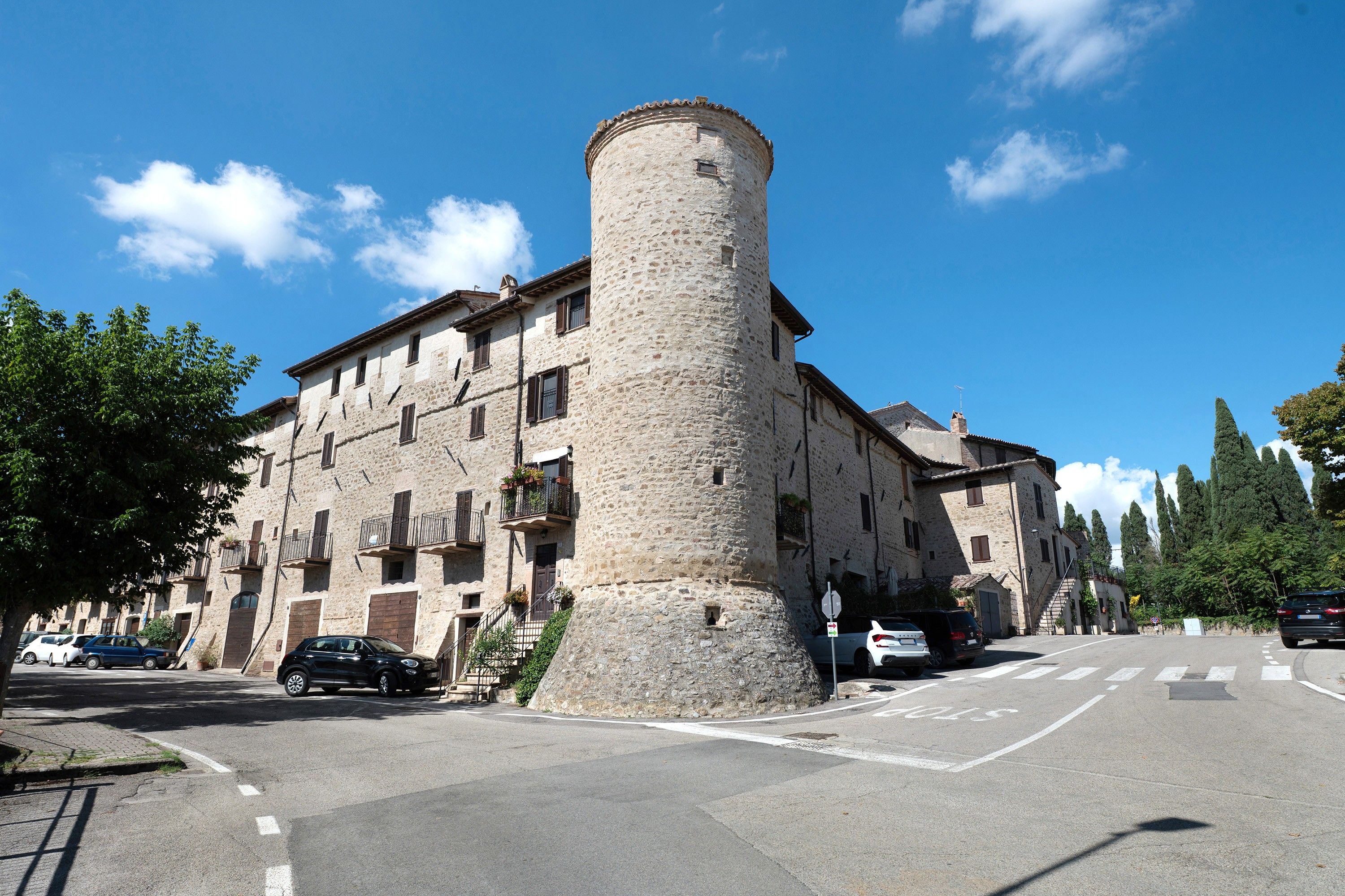 Un edificio storico in pietra con una torre imponente, una strada antistante e auto parcheggiate.