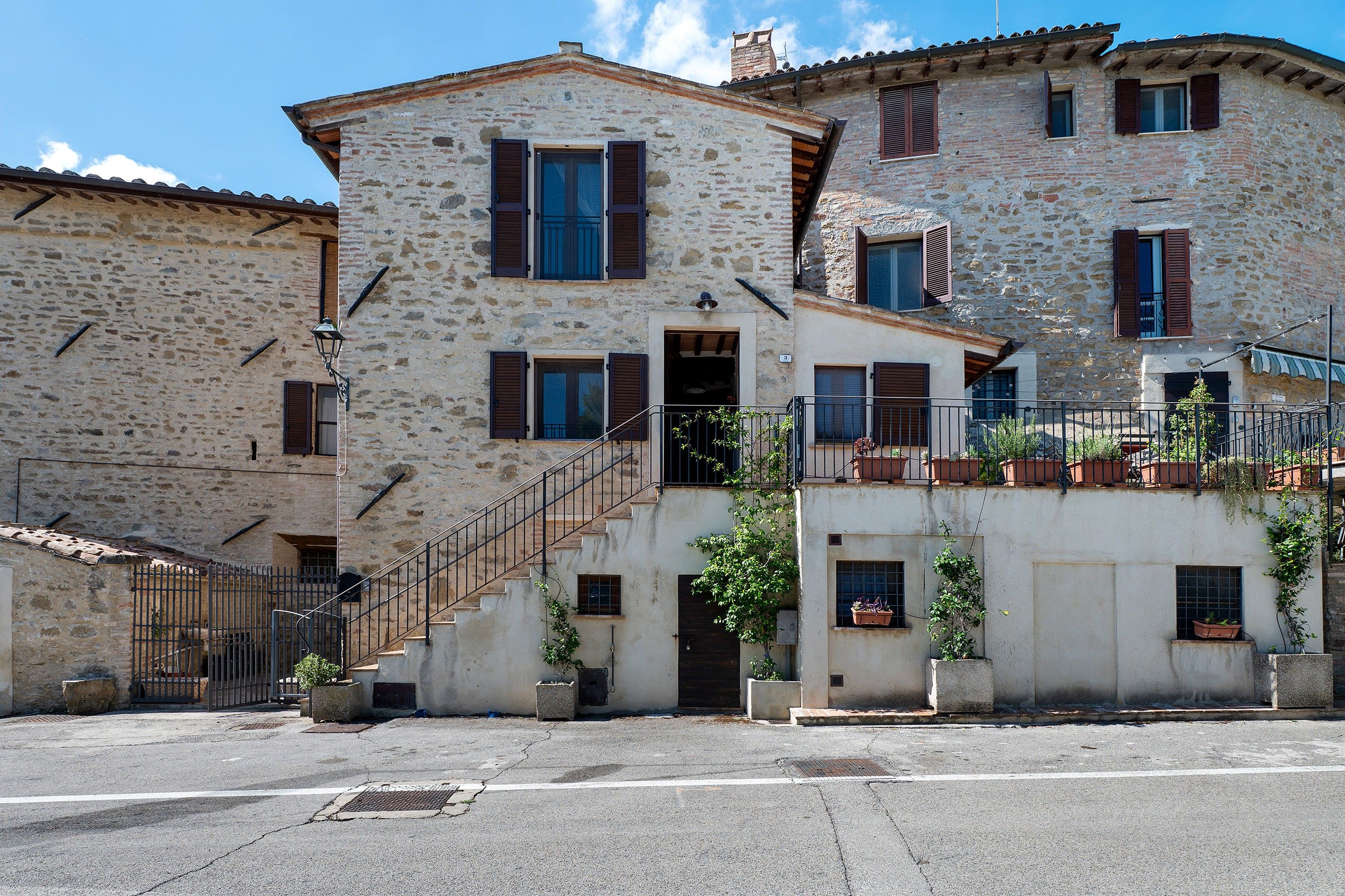 Un bâtiment en pierre de deux étages, avec un escalier menant à l’entrée, des plantes en pot et une rambarde métallique à l’étage supérieur.