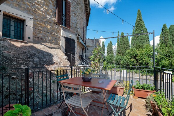A balcony with a table and chairs, potted plants, and a stone building in the background.