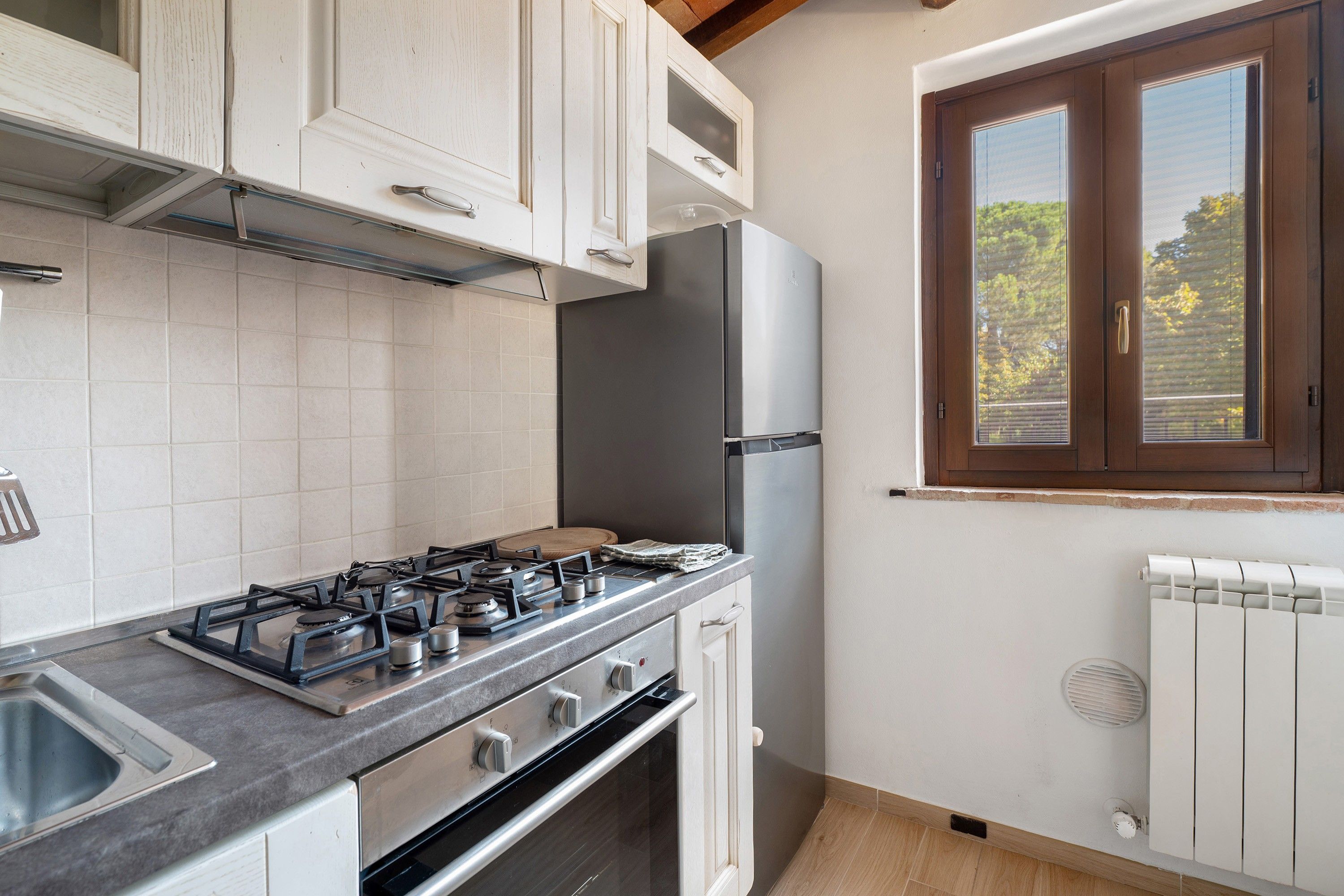 Una cocina con armarios blancos, cocina a gas, refrigerador y ventana con persianas.