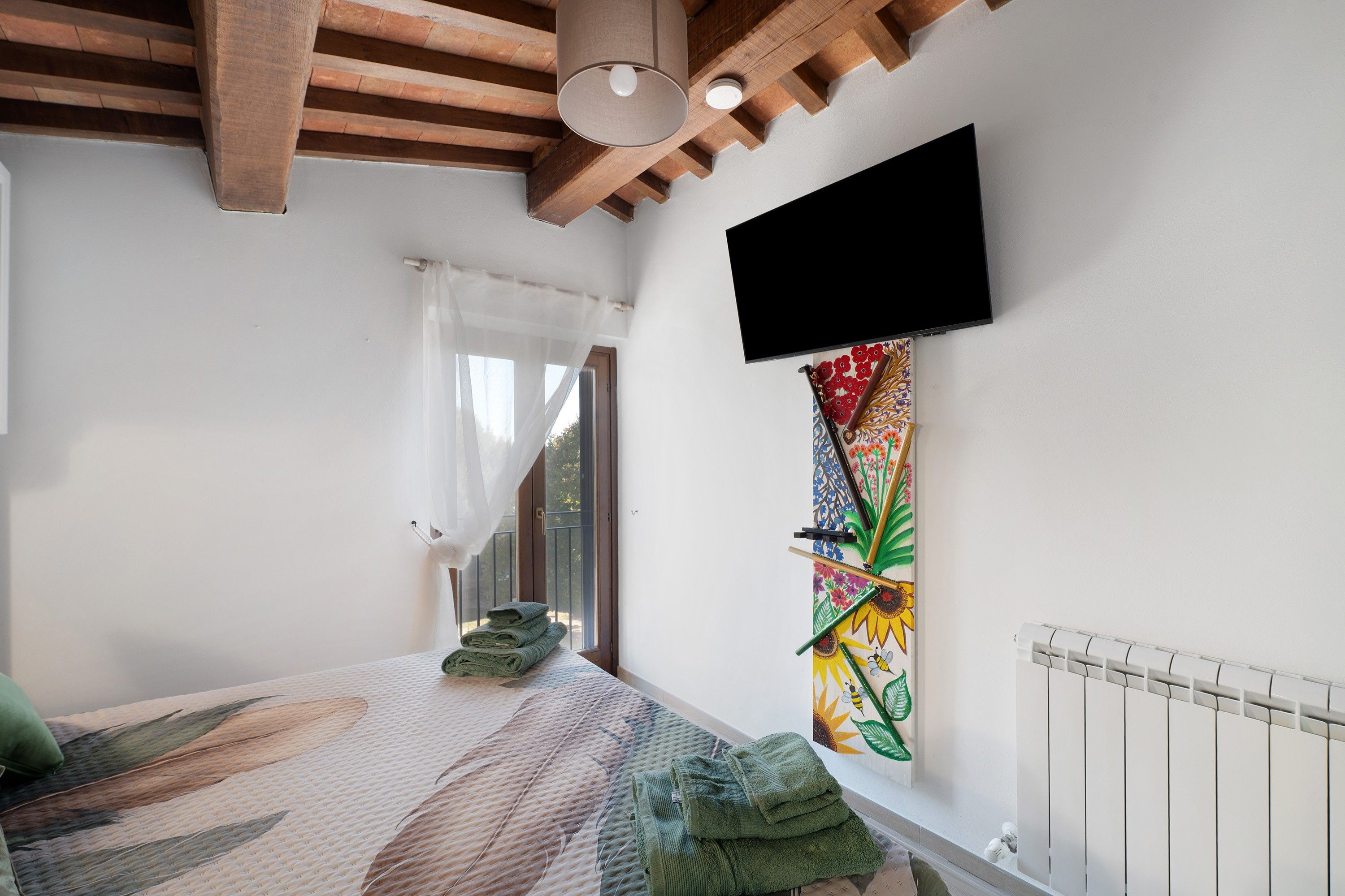 A bedroom with a bed, a flat-screen TV mounted on the wall, a radiator, and a window with a view of trees.