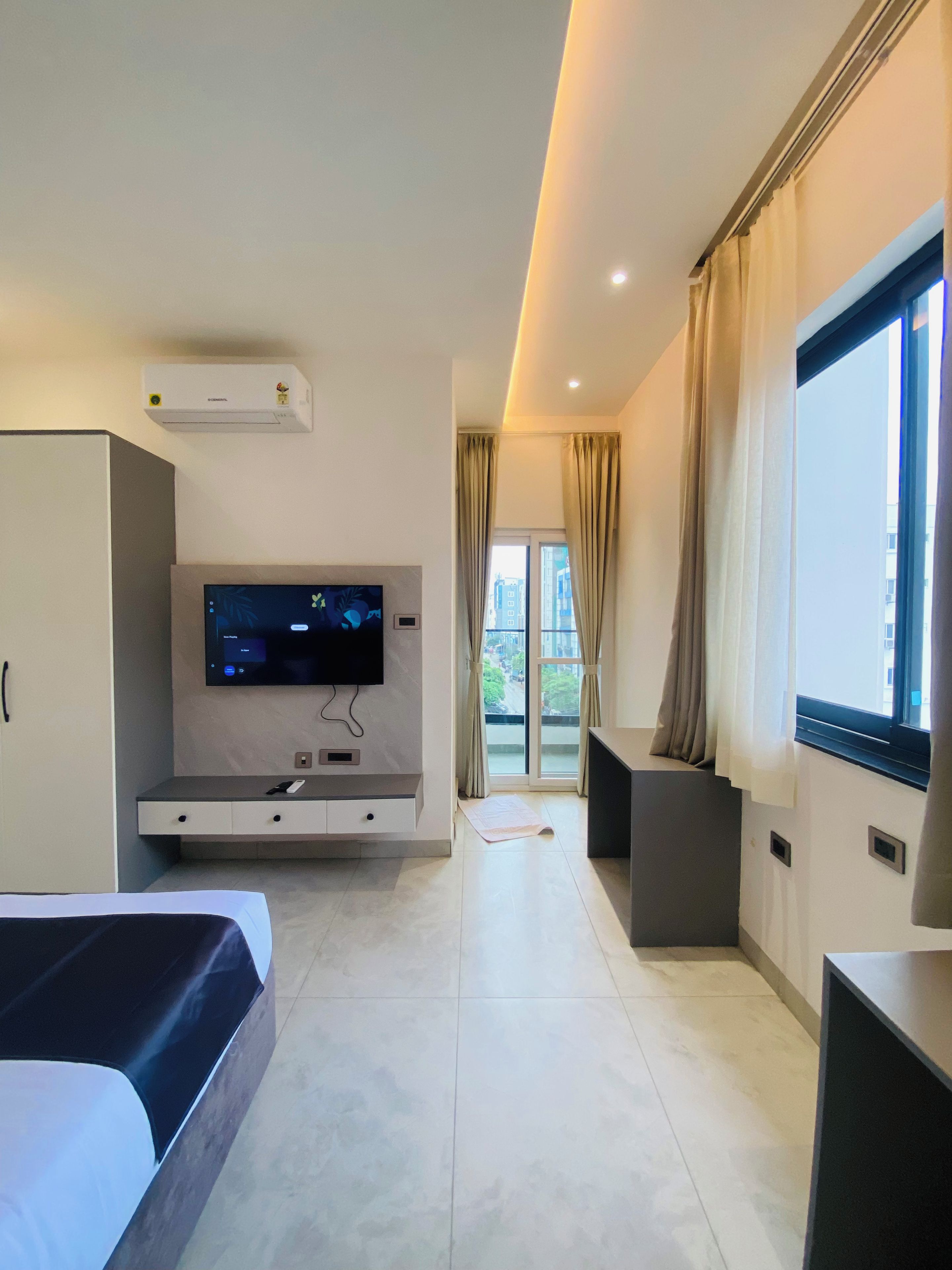 Una habitación de hotel moderna con una cama, un televisor de pantalla plana montado en la pared, un escritorio con una silla y un ventanal con cortinas.