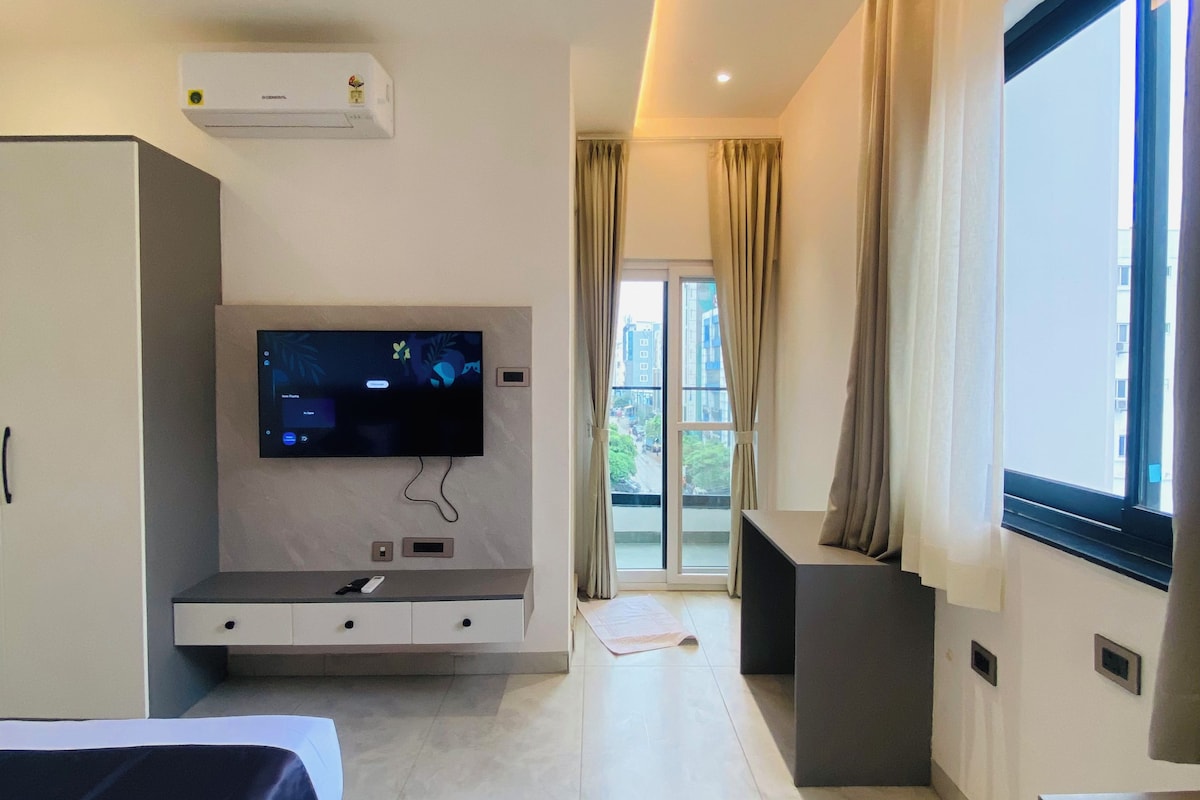 A modern hotel room with a bed, a flat-screen TV mounted on the wall, a desk with a chair, and a large window with curtains.