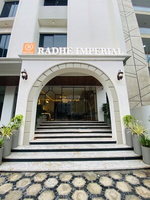 La entrada del Hotel Radhe Imperial, con un diseño moderno, presenta un amplio arco de entrada, escalones que conducen a la recepción y plantas en macetas a ambos lados.