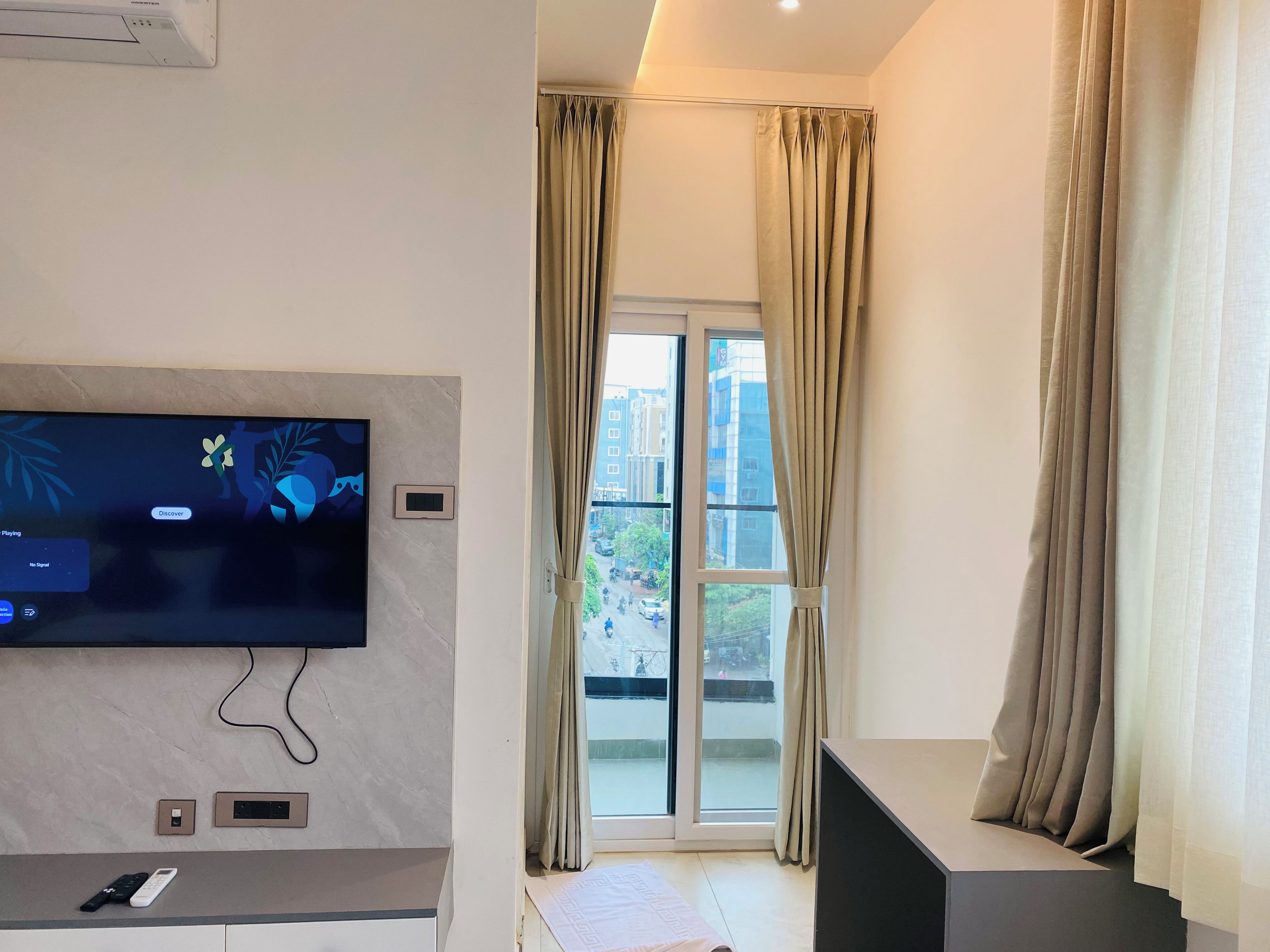A modern living room with a flat-screen TV mounted on the wall, a remote control on a counter, and a sliding glass door leading to an outdoor area.