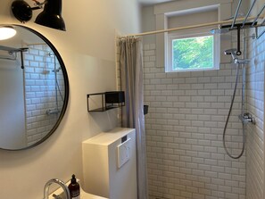 Ein Badezimmer mit einer weißen Duschkabine, einem Fenster mit Blick auf Grünflächen, einem runden Spiegel und einer Seifenspender.