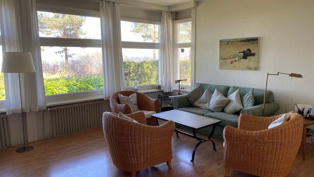 Sala de estar com sofá verde, cadeiras de vime, mesa de centro e janelas amplas com cortinas.