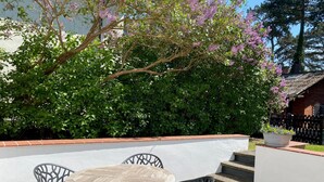 Um pátio externo com mesa redonda e quatro cadeiras, cercado por um muro baixo branco e uma árvore florida.